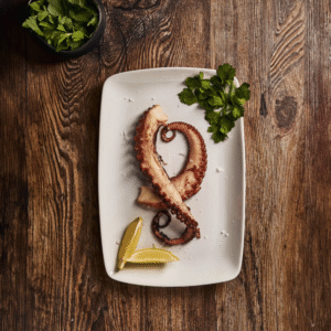 Grilled octopus tentacle on a white plate with lemon wedges and parsley on a wooden table.