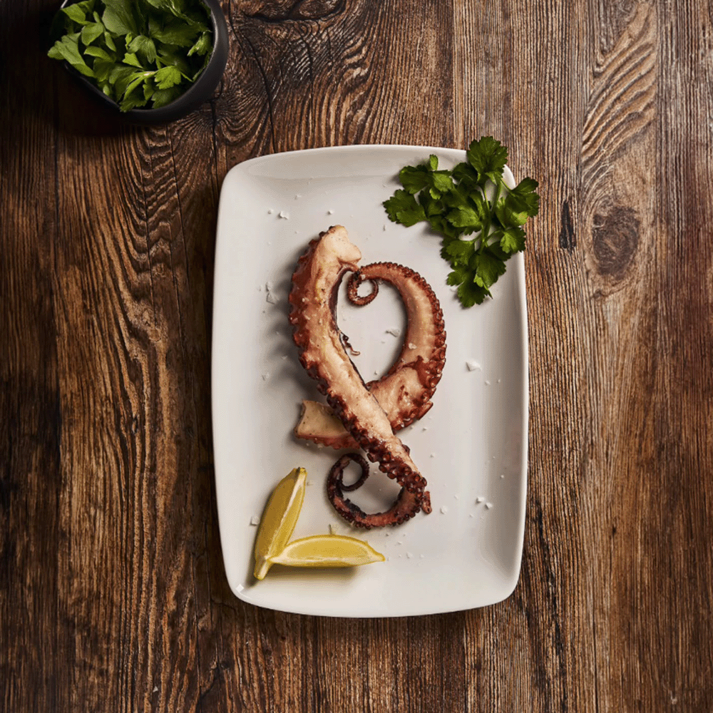 Grilled octopus tentacle on a white plate with lemon wedges and parsley on a wooden table.