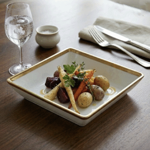 Plate of roasted root vegetables (carrot, parsnip, potatoes, beets) garnished with herbs on a white square dish with a gold rim, beside a glass of sparkling water and cutlery on a wooden table