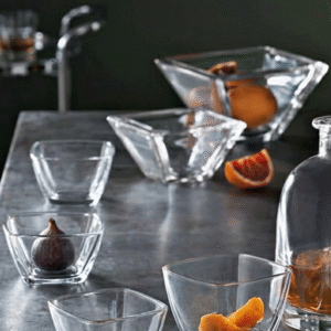 Assorted clear glass bowls and a bottle of amber liquor on a dark bar counter, with orange wedges and a fig inside bowls.