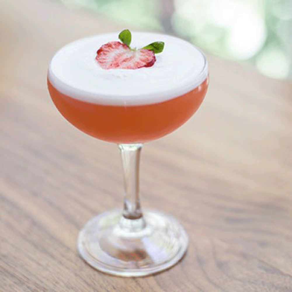 Pink cocktail in a coupe glass with a frothy top, garnished with a strawberry slice and mint leaves on a wooden table.