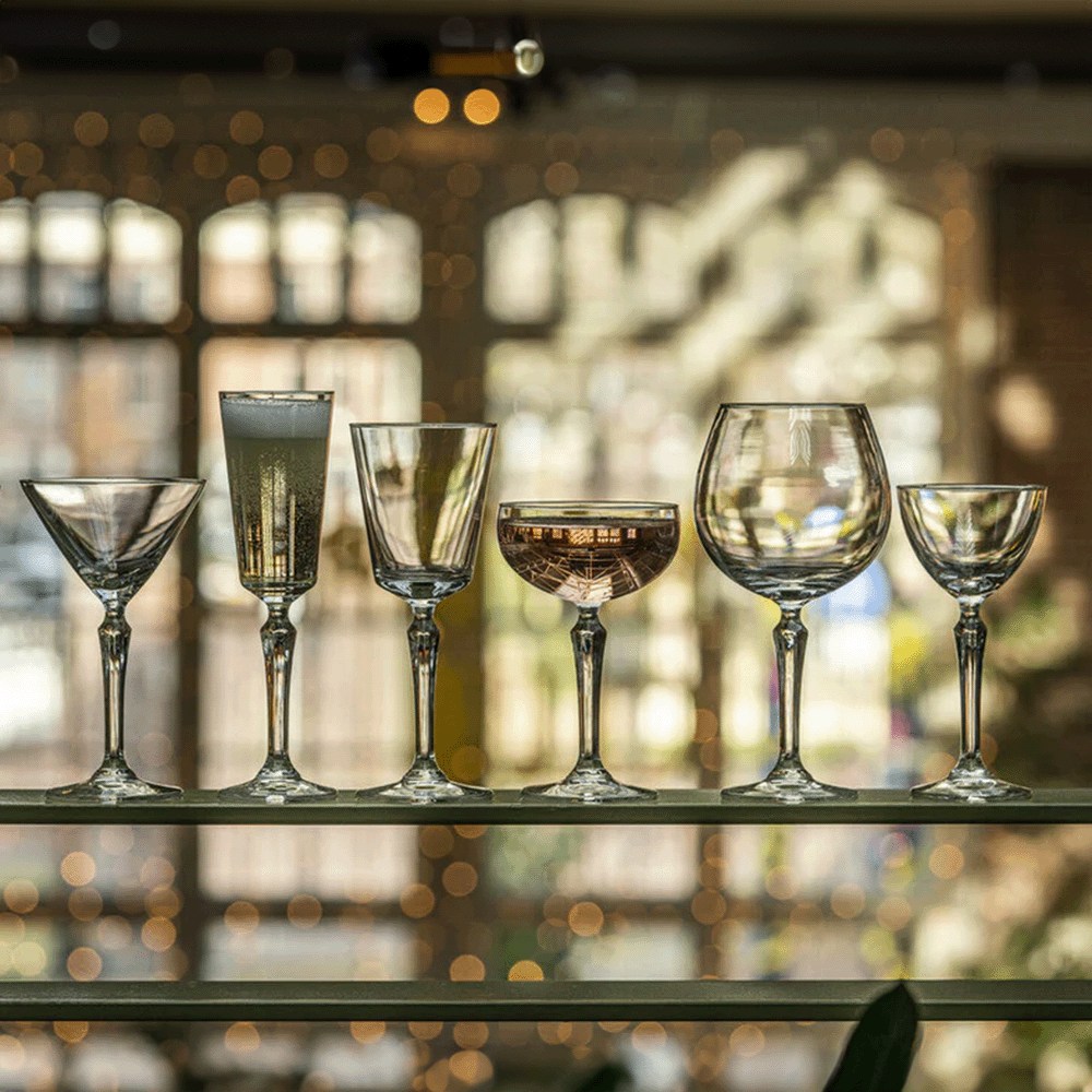 Seven different wine and cocktail glasses lined up on a glass shelf against a blurred, bokeh-filled bar background.