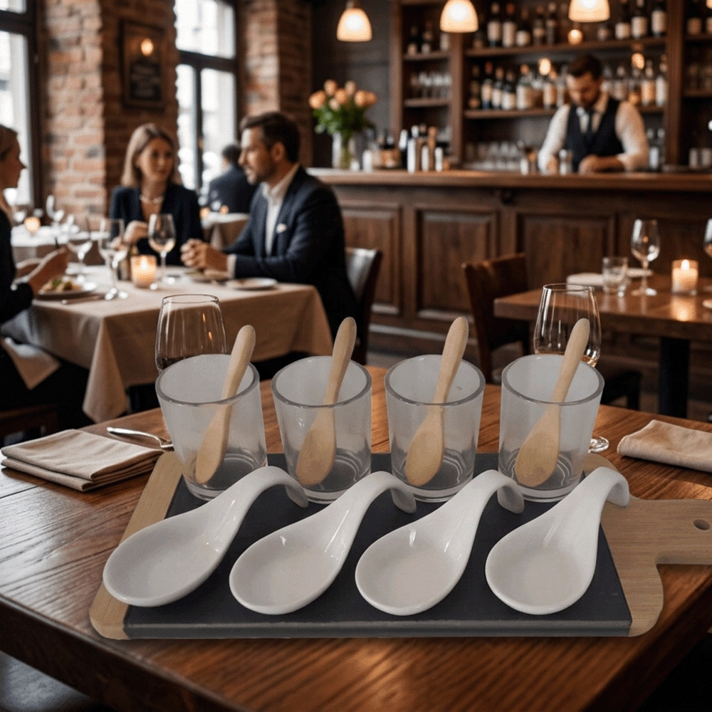 A restaurant table with a four-piece tasting set: four glass cups each with a wooden spoon, and four white ceramic spoons on a tray in the foreground, diners seated in the background.