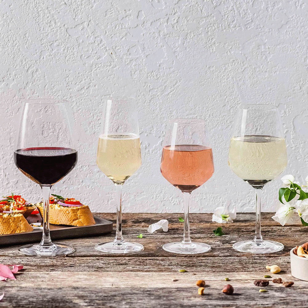 Four wine glasses with red, white, rosé, and light white wine on a weathered wooden table; tapas and white flowers nearby.