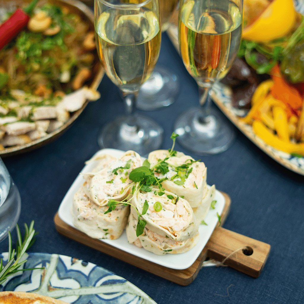 Plate of herbed cream cheese wraps garnished with greens on a small white dish, on a dark table with champagne flutes in the background.