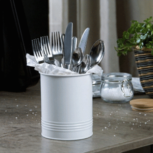 Metal cutlery—forks, knives, and spoons—stuck in a white cylindrical holder lined with tissue on a wooden counter with a salt jar and plant in the background.