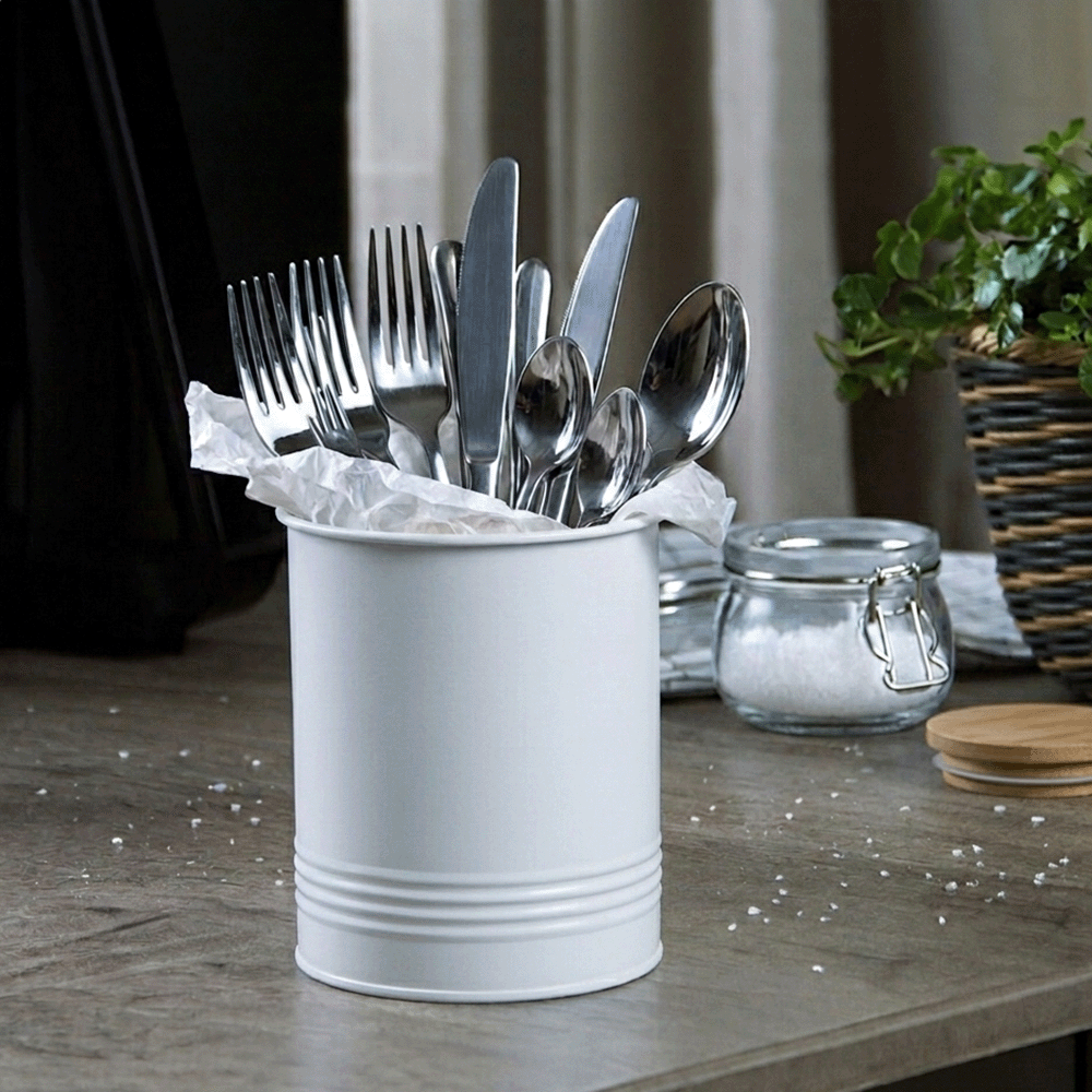 Metal cutlery—forks, knives, and spoons—stuck in a white cylindrical holder lined with tissue on a wooden counter with a salt jar and plant in the background.