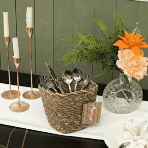 Decorative table setting: woven basket holding forks and spoons, with copper candle holders and a floral centerpiece against a green wooden wall.