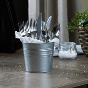 Metal bucket filled with forks, knives, and spoons wrapped in tissue on a wooden table, with a sugar jar and plant in the background.