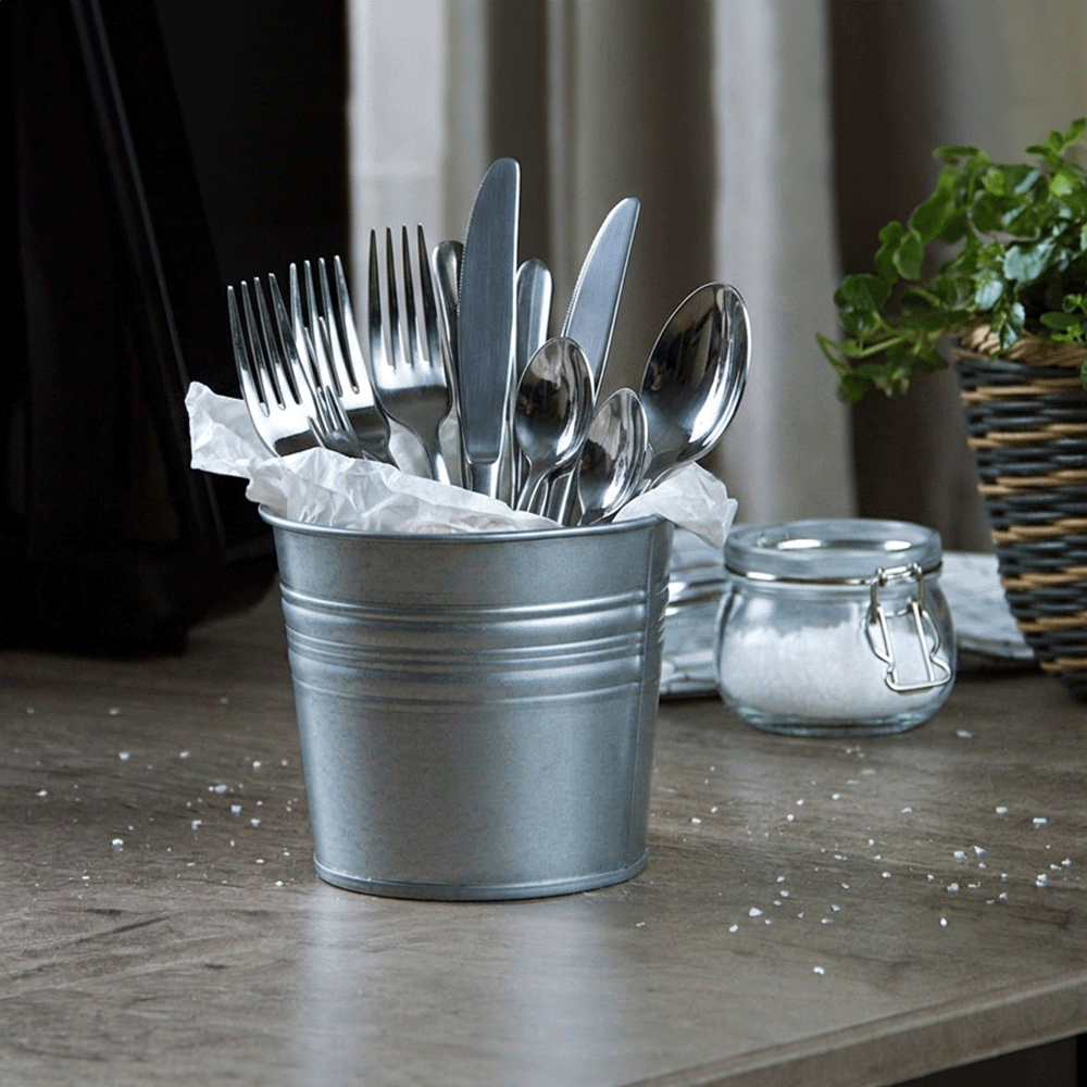 Metal bucket filled with forks, knives, and spoons wrapped in tissue on a wooden table, with a sugar jar and plant in the background.