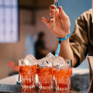 Two crystal glasses filled with orange punch and large ice cubes on a bar, a blurred person with a blue wristband in the background.