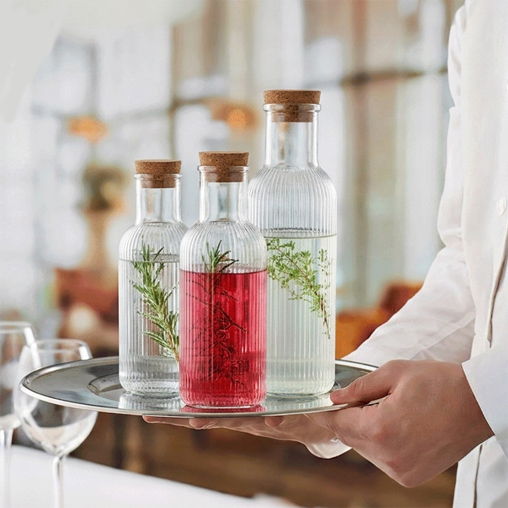 Three glass bottles with herb sprigs on a silver tray, carried by a person in a white coat; the middle bottle contains red infusion.