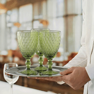 Three emerald-green cut-glass goblets resting on a silver tray carried by a server in a restaurant setting.