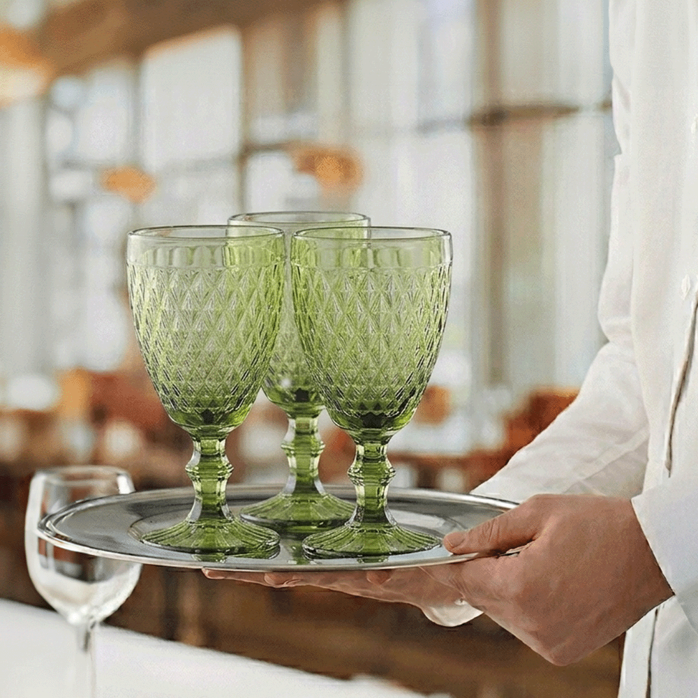 Three emerald-green cut-glass goblets resting on a silver tray carried by a server in a restaurant setting.