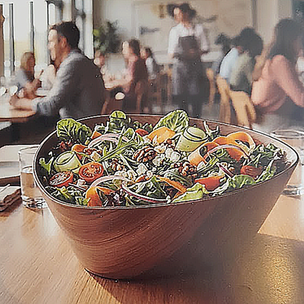 Large wooden bowl of colorful mixed salad on a restaurant table with diners in the background.