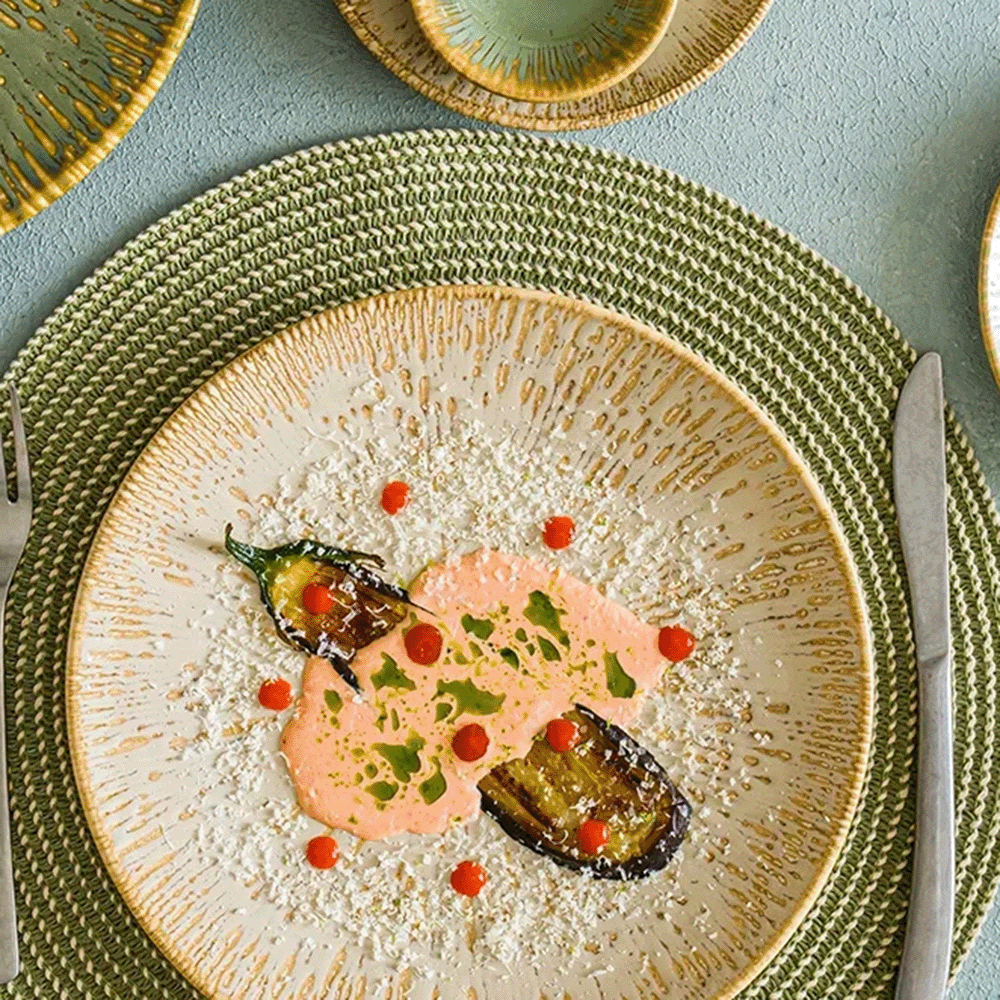 Gourmet plate with pink sauce, roasted eggplants, green herbs, and red sauce dots on a speckled white plate set on a green woven placemat, with cutlery nearby.