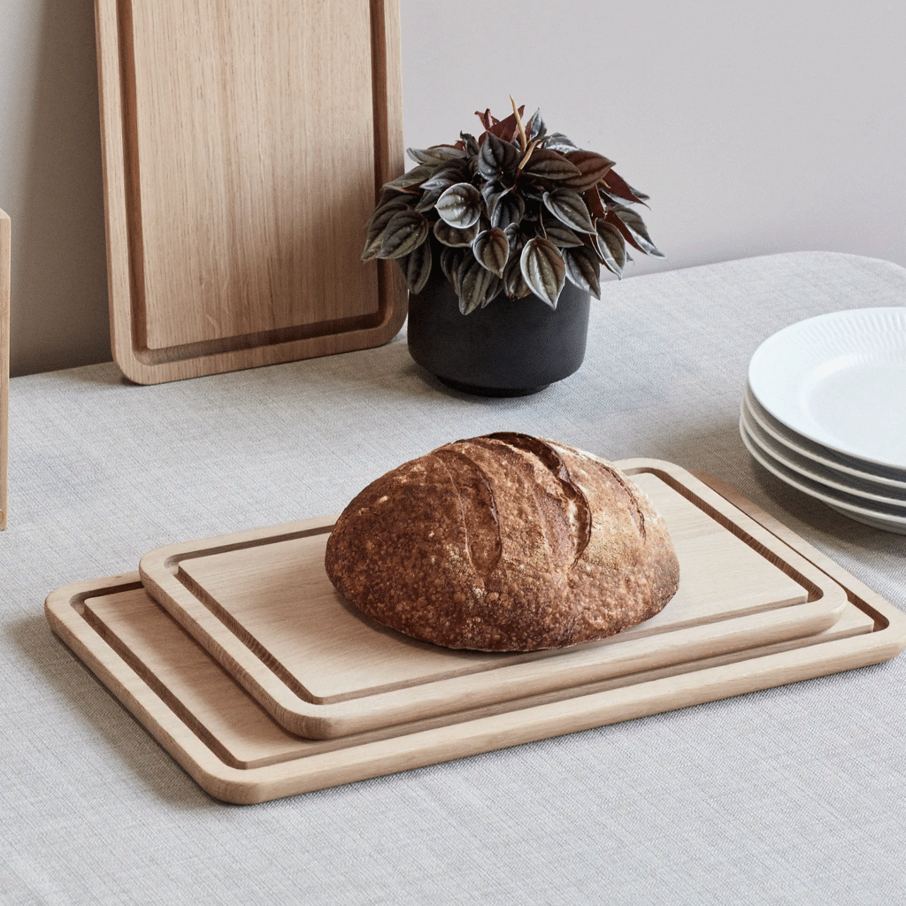 Crusty round loaf on a light wooden cutting board on a dining table, with a leafy potted plant and white plates nearby.