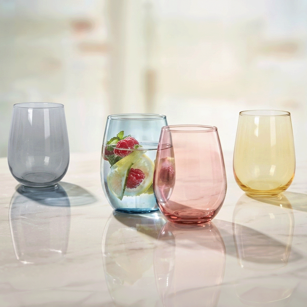 Four colorful stemless glasses on a reflective marble table; blue glass with fruit infusion, pink and yellow glasses nearby, gray glass to the left.
