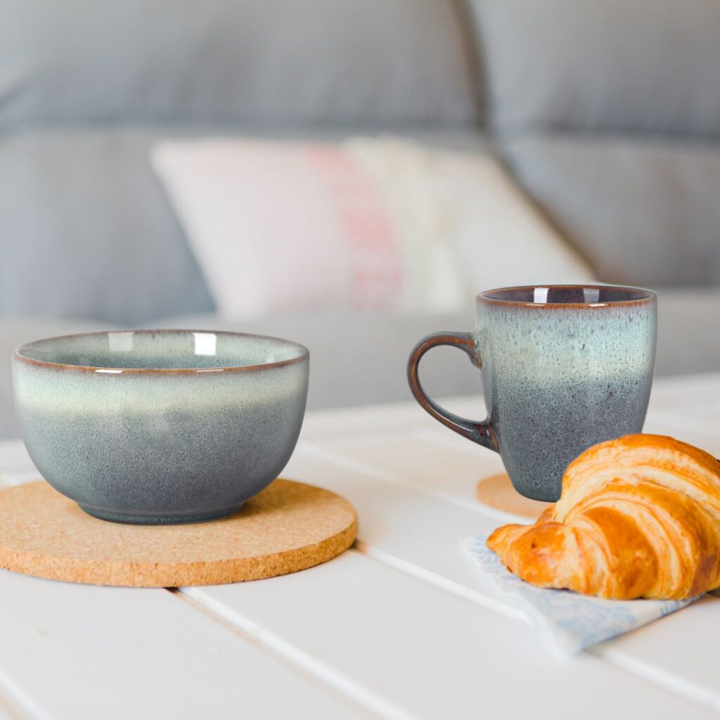 Two blue-gray ceramic mugs on cork coasters with a croissant on a napkin on a white table, soft couch in the background.