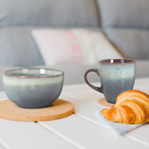 Two blue-gray ceramic mugs on cork coasters with a croissant on a napkin on a white table, soft couch in the background.