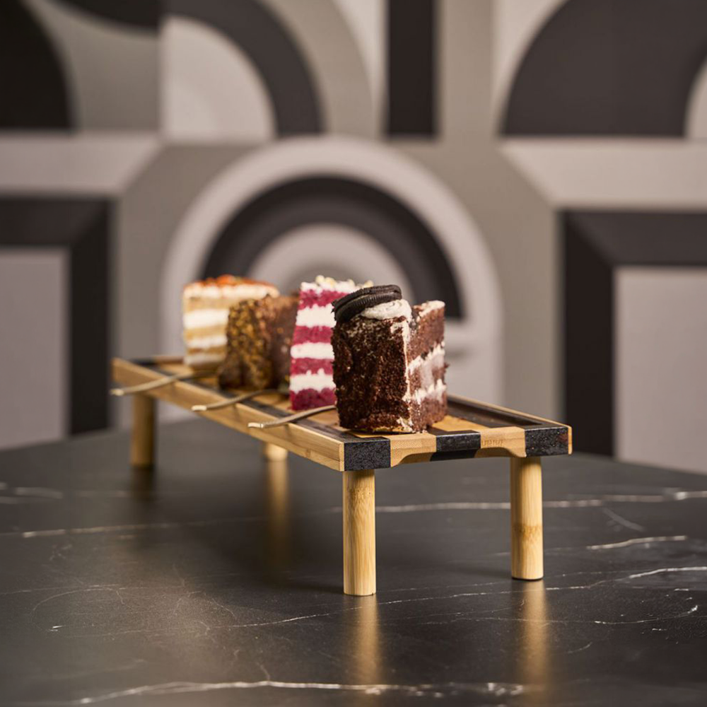 Assorted mini cake slices displayed on a small wooden elevated stand against a geometric backdrop.