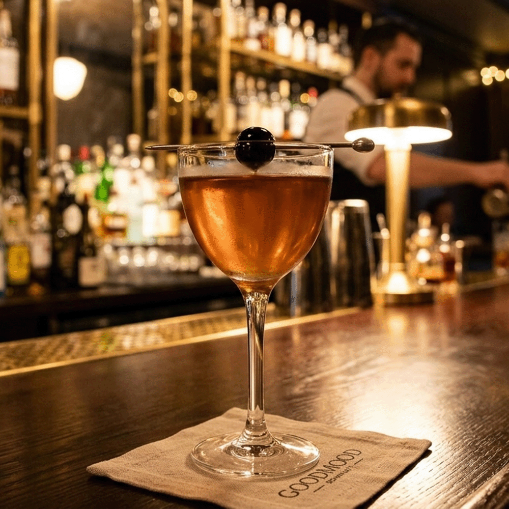 Amber cocktail in a coupe glass with a black olive on a skewer resting across the rim; bar shelves blurred in the background, napkin on the counter.