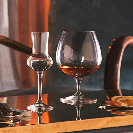 Two crystal glasses on a brass bar, one filled with whiskey and the other with a clear drink, with lemon slices on a plate in the background.