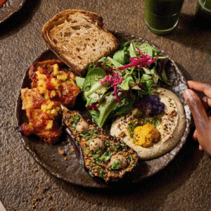 Plate with hummus, roasted eggplant, greens, bread, and diced tomato relish; a hand reaches in from the right.