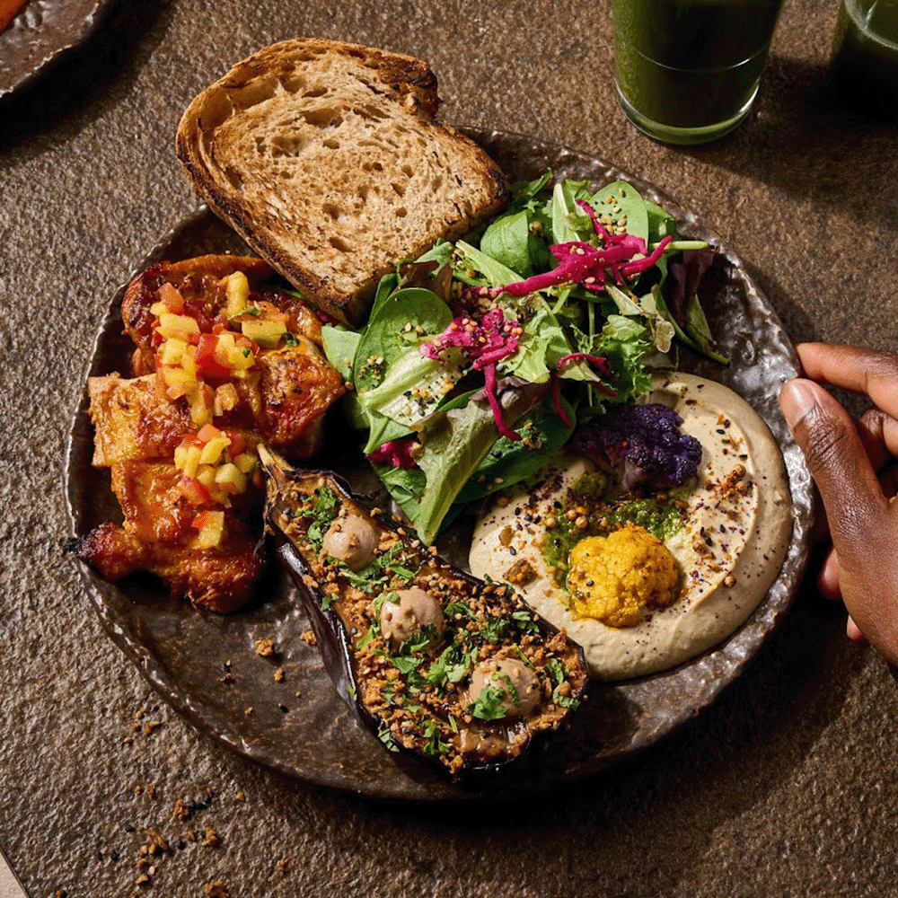 Plate with hummus, roasted eggplant, greens, bread, and diced tomato relish; a hand reaches in from the right.