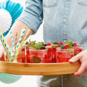 Person holding a wooden tray with red berry drinks garnished with mint, berries, and colorful straws on a bright background
