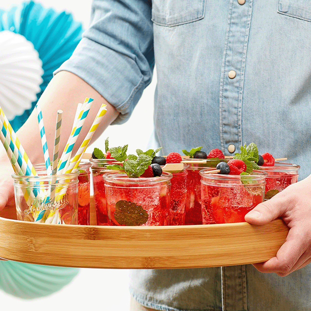 Person holding a wooden tray with red berry drinks garnished with mint, berries, and colorful straws on a bright background