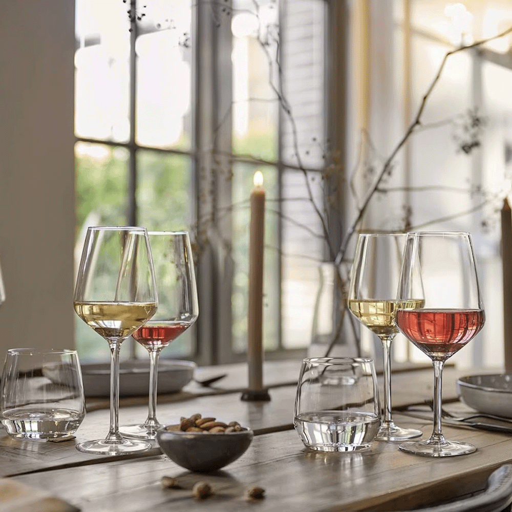 Elegant dining table set with white and rosé wine glasses, a lit candle, and a bowl of nuts by a sunlit window.