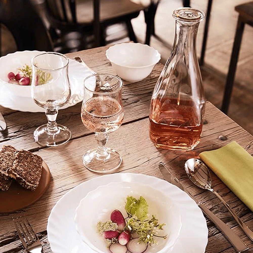 Rustic dining table set with white plates, a rosé decanter, two wine glasses, bread slices, and a small salad bowl.