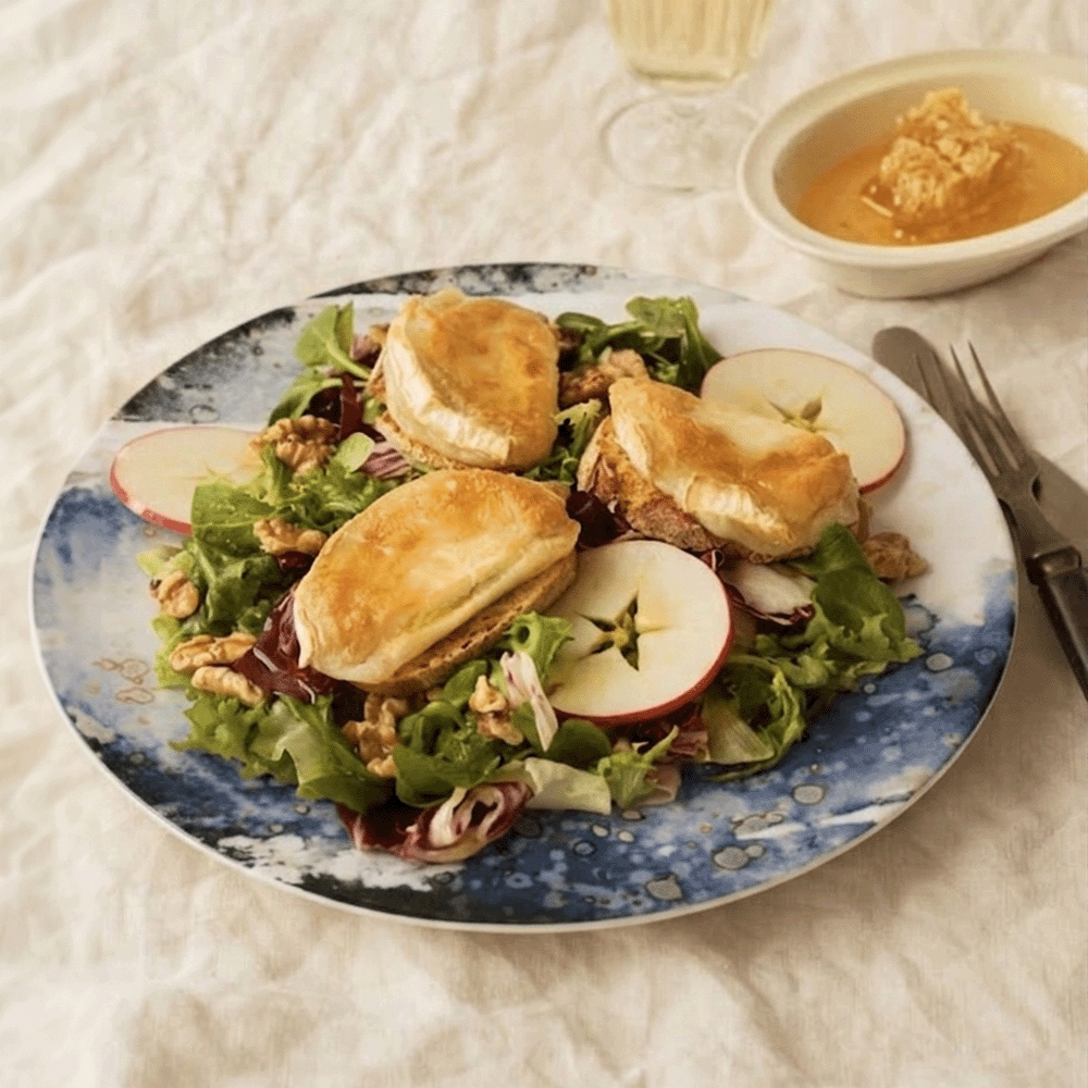 Plate of mixed greens with small croissant sandwiches, apple slices, and walnuts; a bowl of dressing sits nearby on the table.