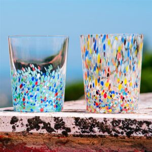 Two clear glasses with multicolored confetti-like dots inside, resting on a weathered ledge against a blue sky.