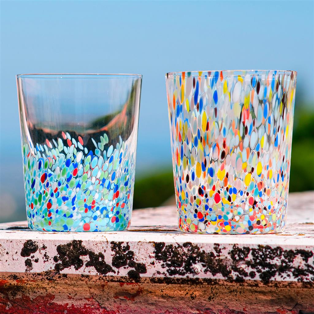 Two clear glasses with multicolored confetti-like dots inside, resting on a weathered ledge against a blue sky.