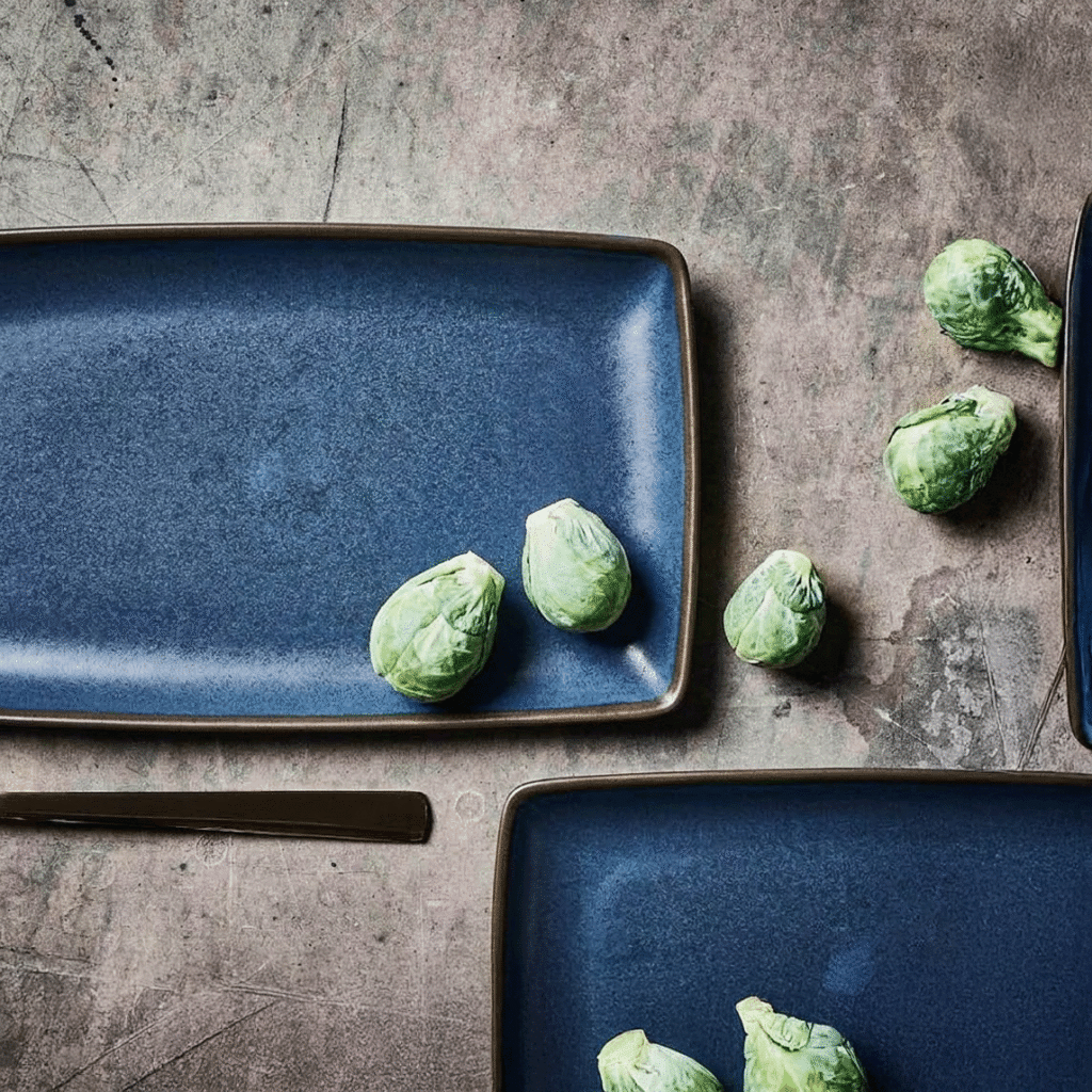 Top-down view of blue ceramic plates with several Brussels sprouts scattered on a textured gray surface